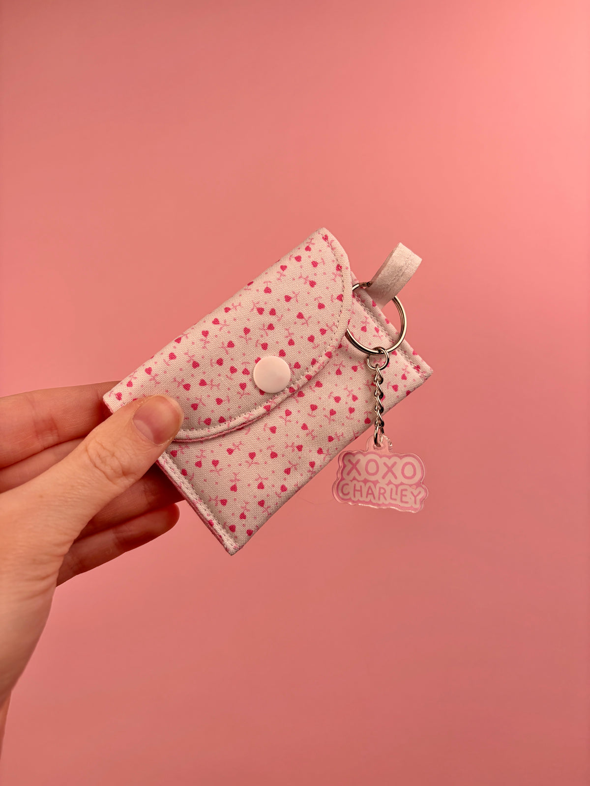 Pink heart-print earbuds case with snap flap, metal ring, and XOXO Charley charm