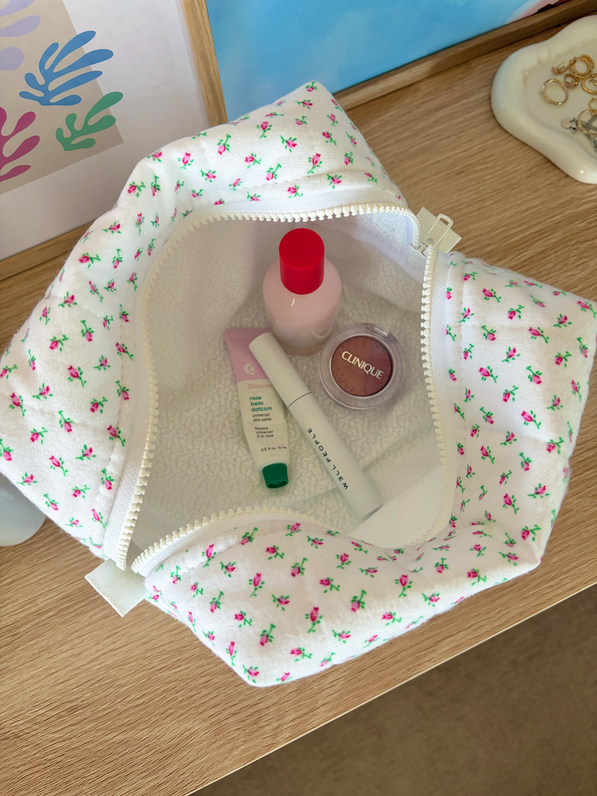 White mini makeup bag with rose floral print, open zip top with visible cosmetics inside.