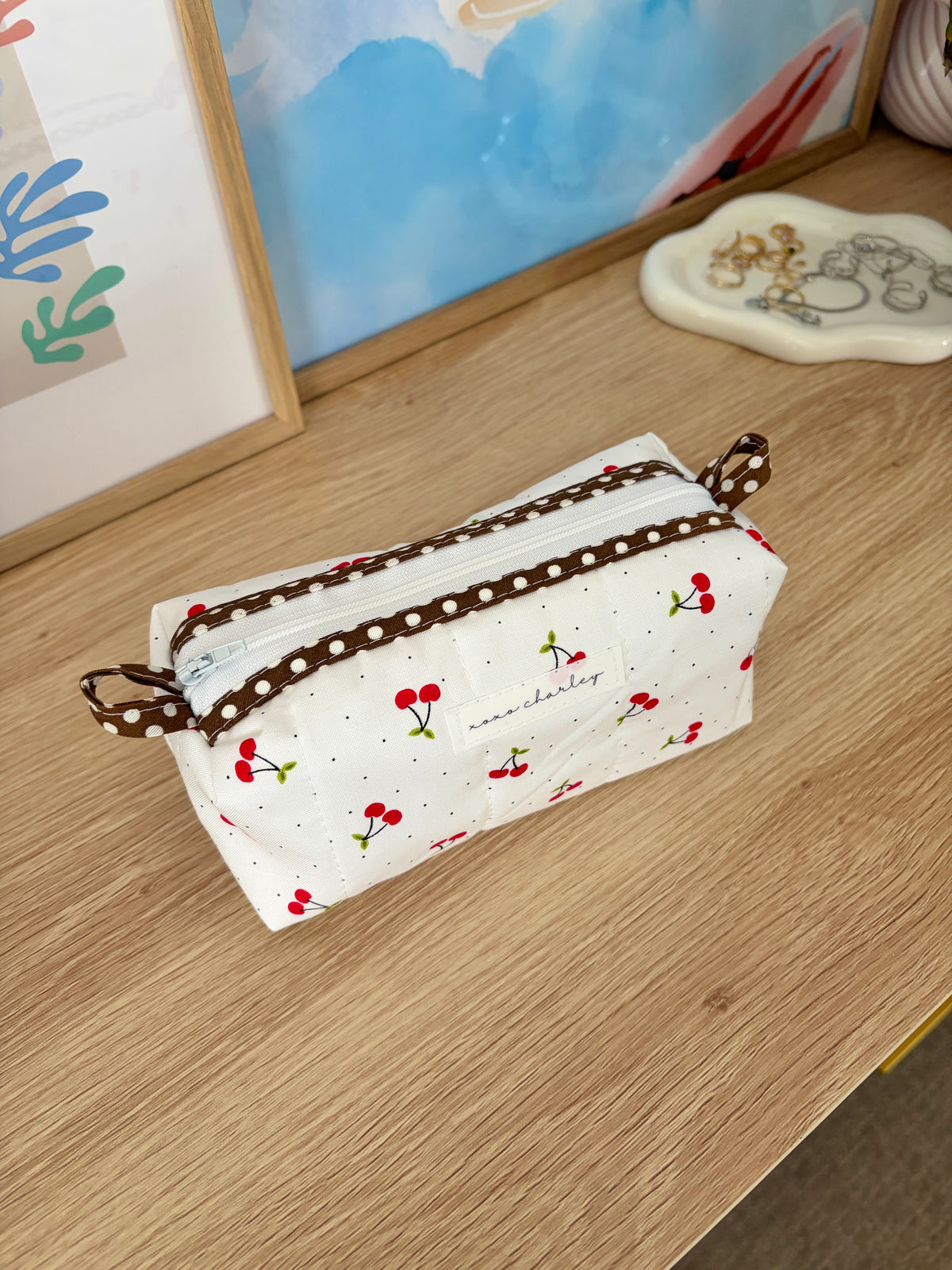 White zippered pouch bag with red cherry print, brown trim, and tied wrist strap
