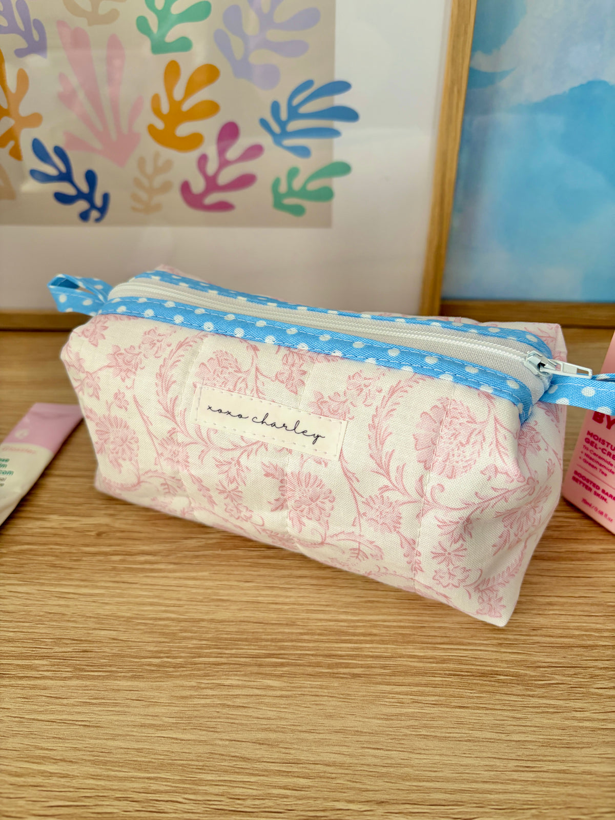 Pink floral zip pouch with blue-trimmed edge and centered brand patch