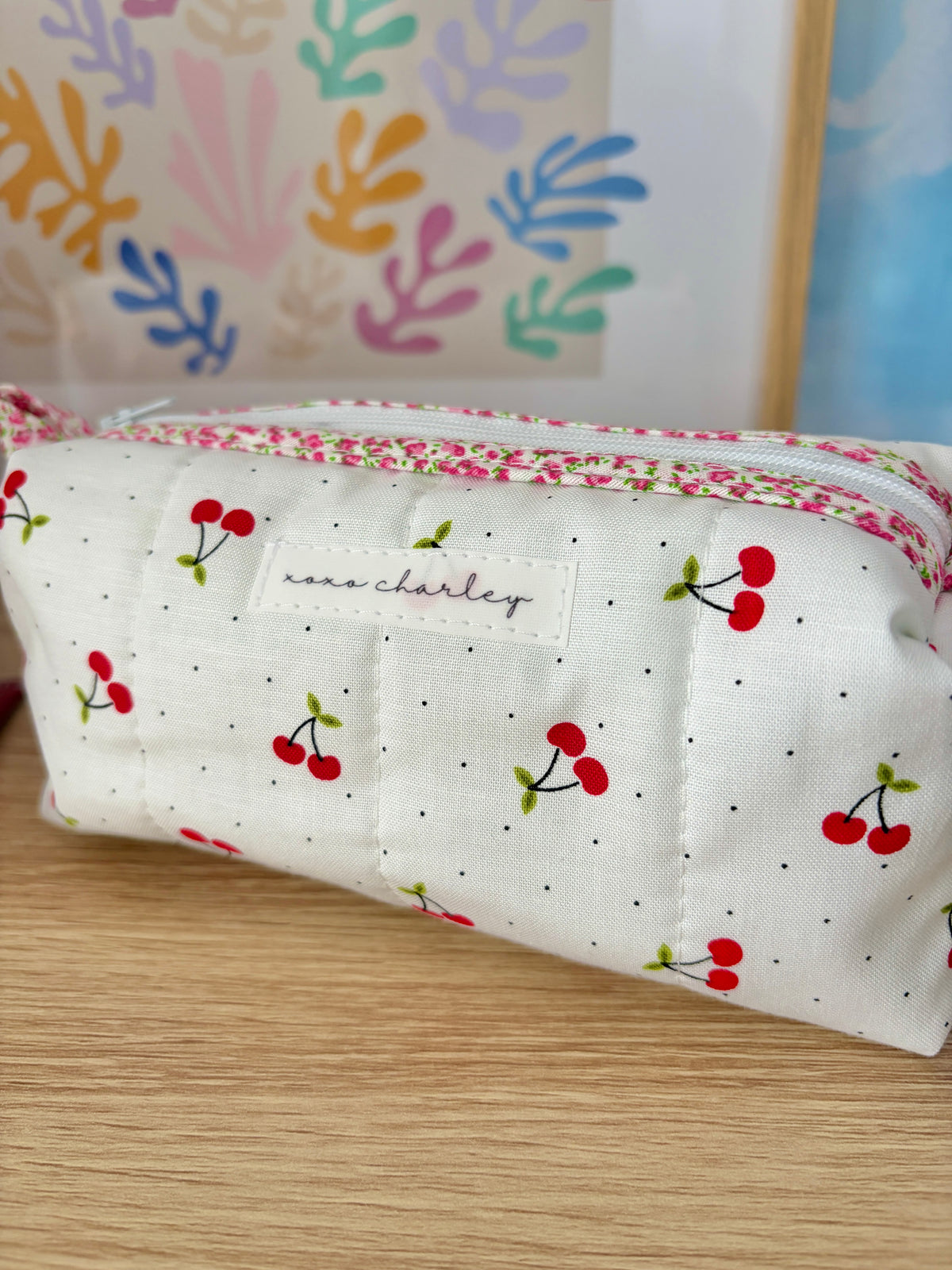 White cherry print zip pouch with logo label patch and pink floral trim; green gingham lining