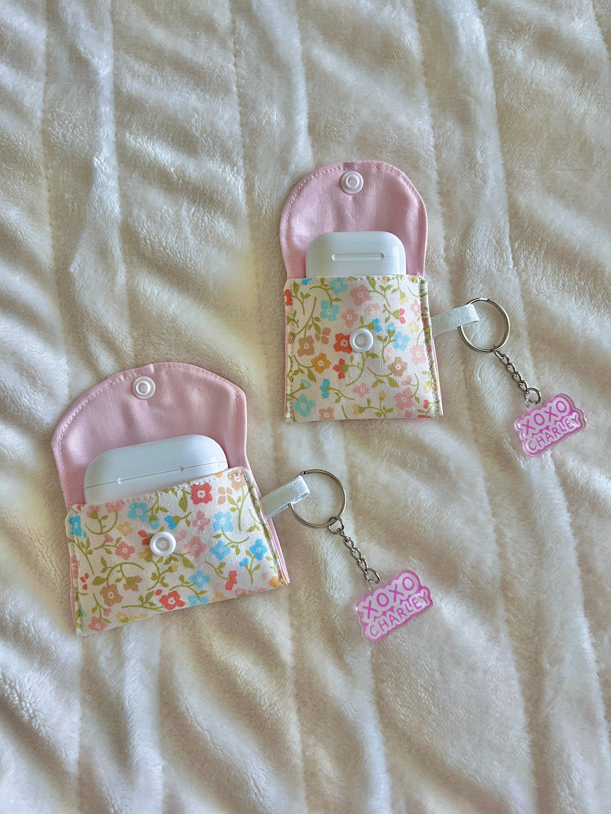 Pastel earbud cases with pink flaps, floral panels, and keychain rings; one holds earbuds