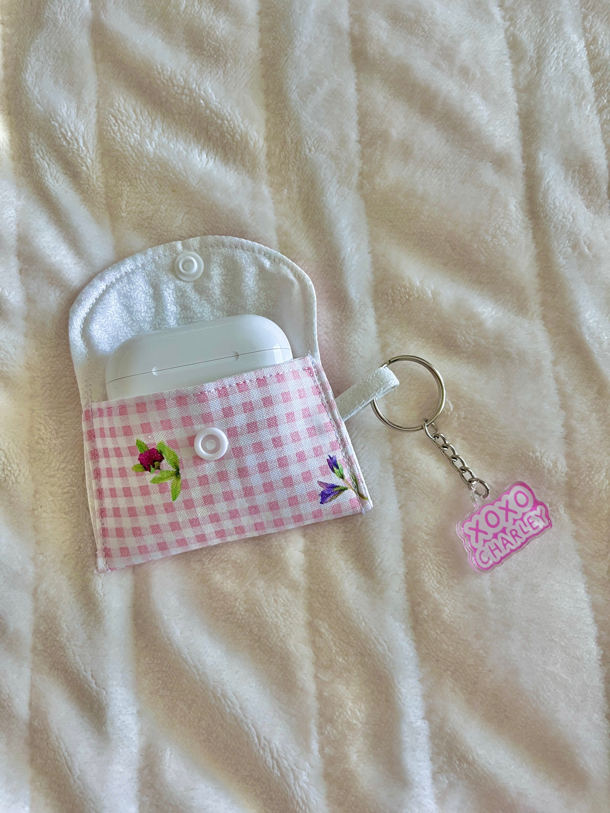 Pink gingham earbuds case with flower embroidery, snap button front, and keychain loop.