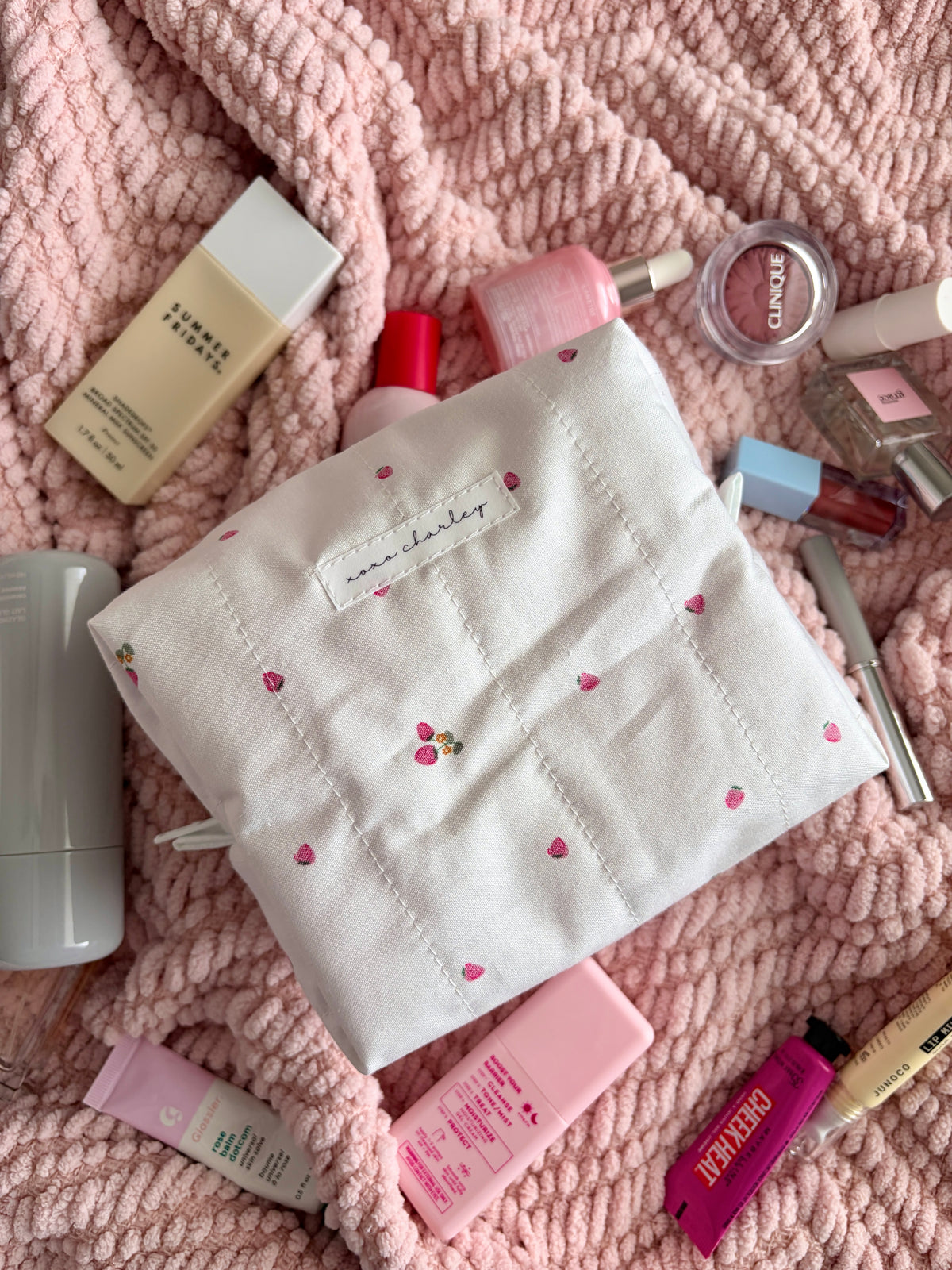 Medium cream makeup bag with embroidered strawberries and stitched logo patch, angled
