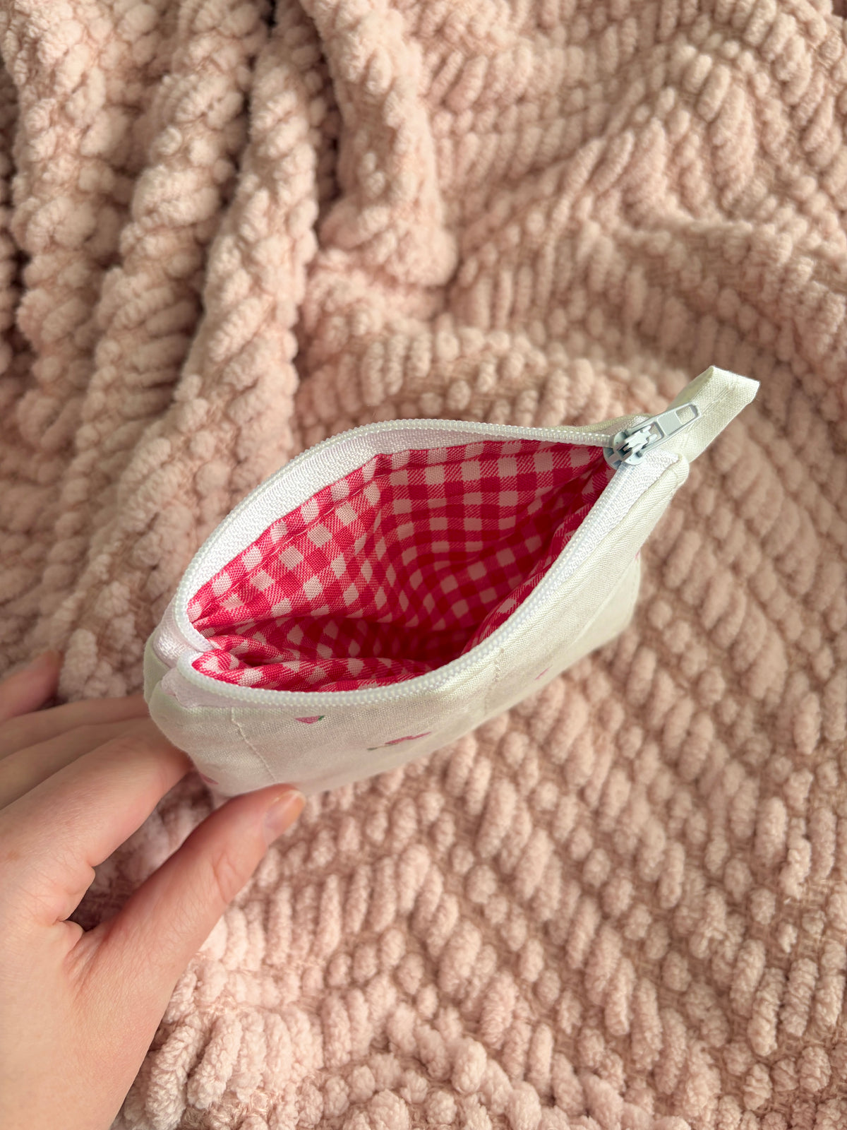 Small white pouch with a red checkered interior held by a hand on a textured pink surface.