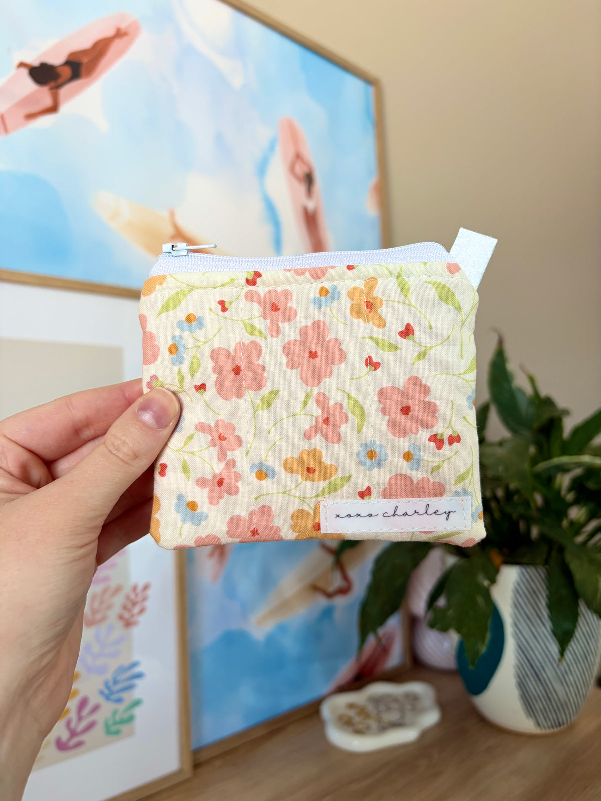 Hand holding a floral-patterned pouch with a plant and framed picture in the background