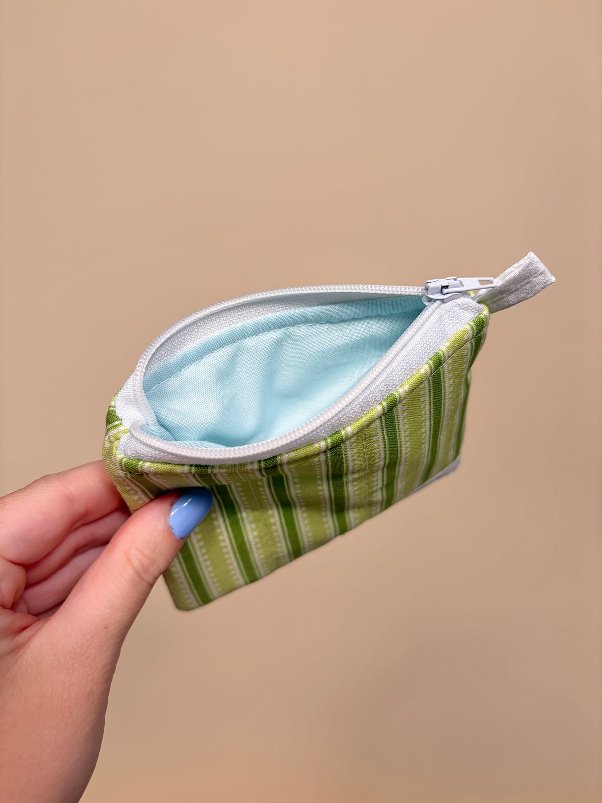 Hand holding a small green striped pouch with a white zipper against a beige background