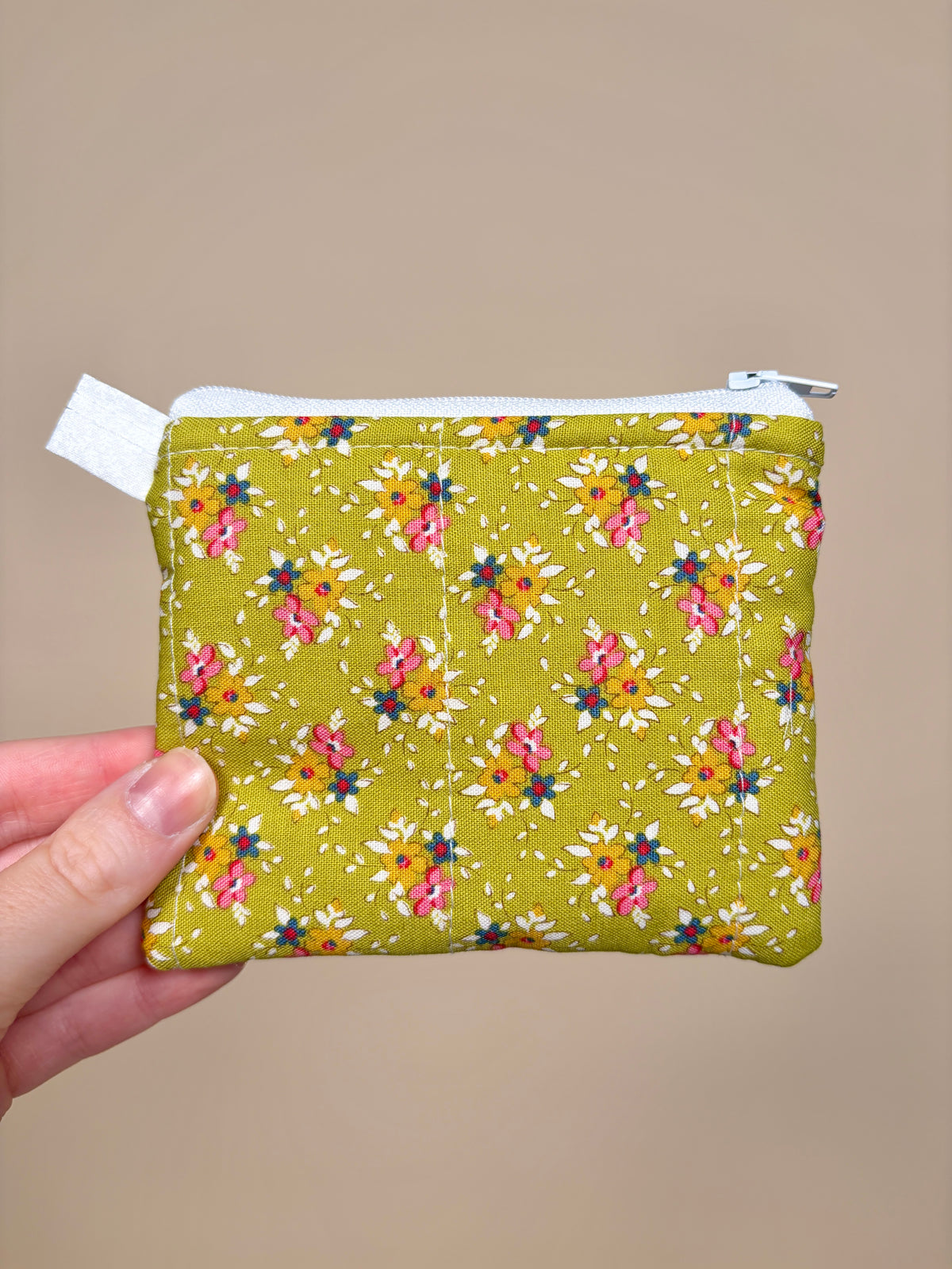 Hand holding a small green fabric pouch with floral pattern against a beige background