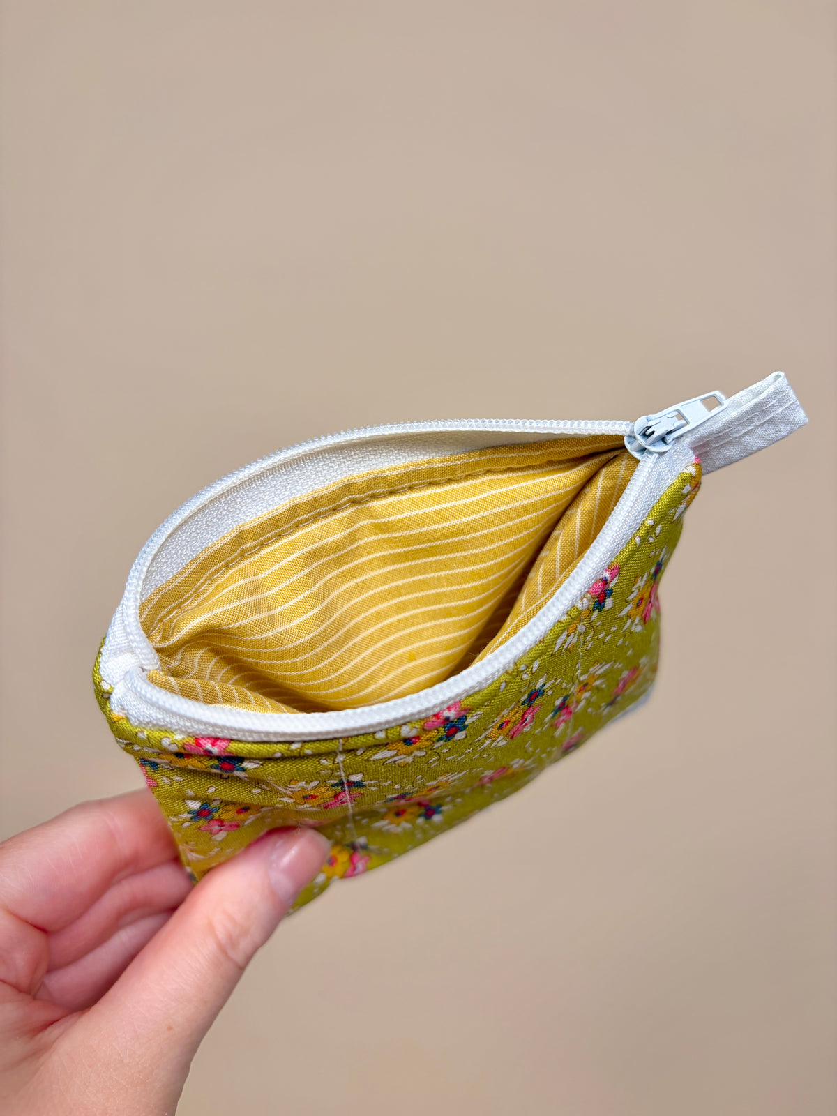 Hand holding a small green fabric pouch with floral pattern and white zipper against a beige background