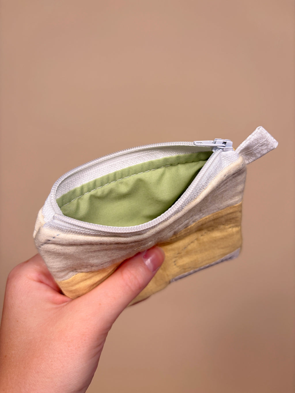 Hand holding a small, round pouch with a zipper against a beige background