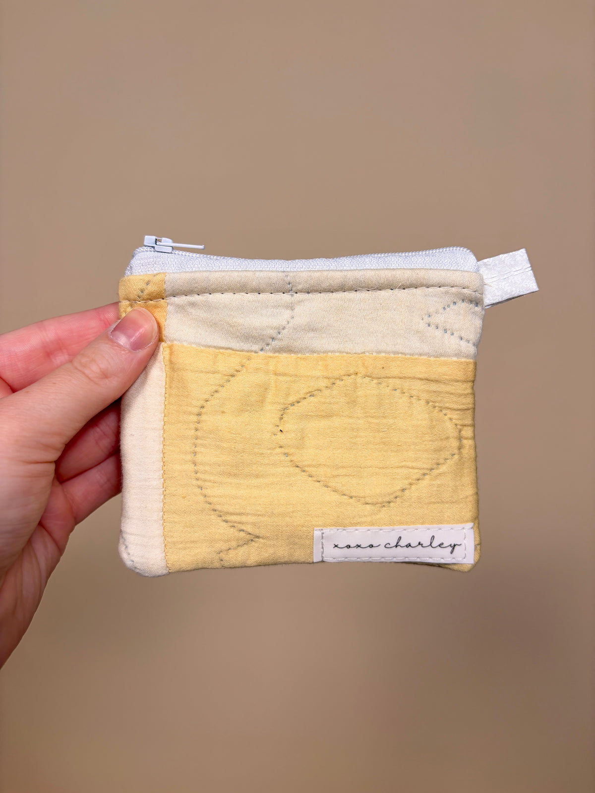 Hand holding a small quilted pouch with a heart design against a beige background