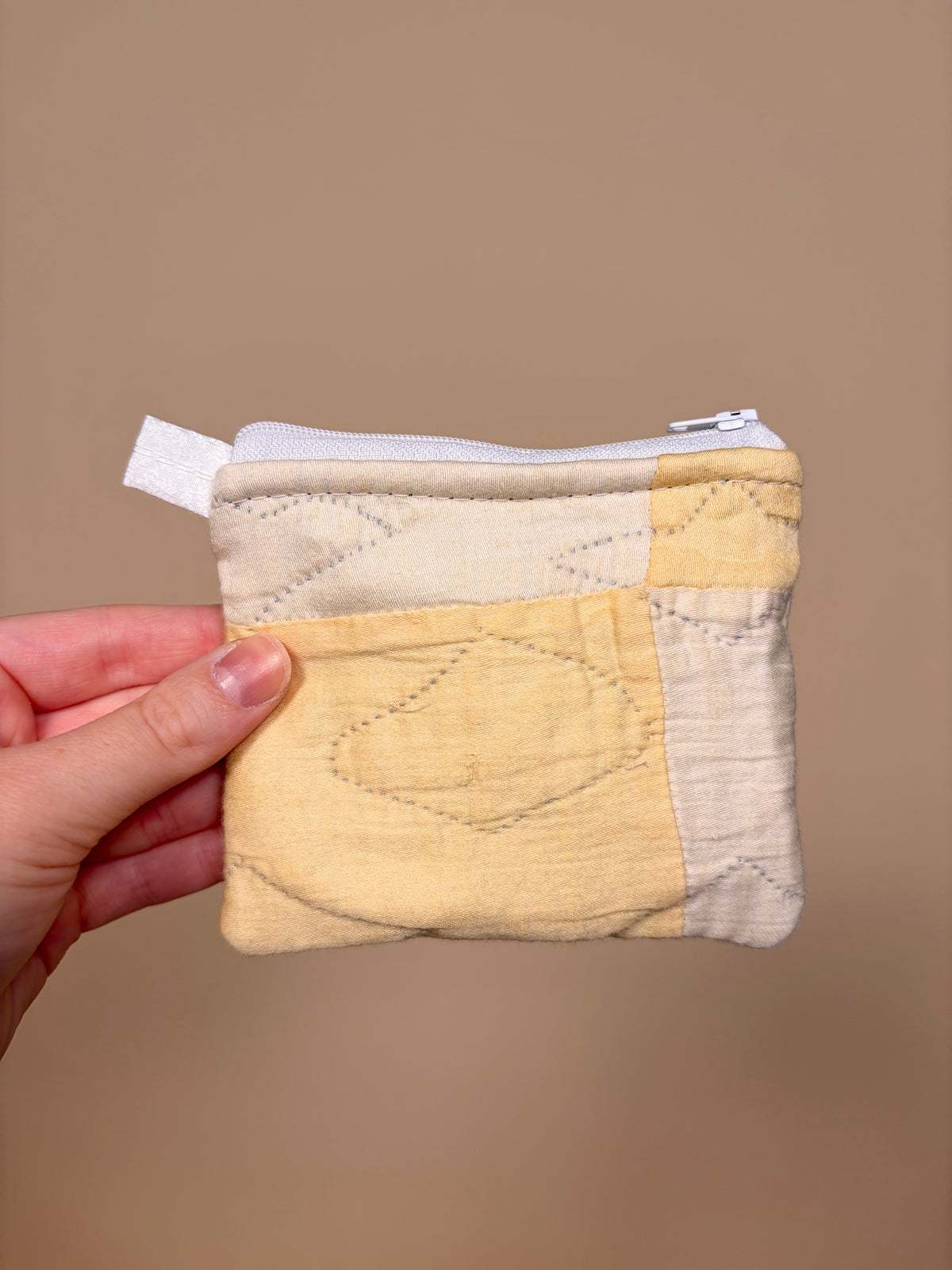 Hand holding a quilted coin purse with a beige and brown design against a brown background