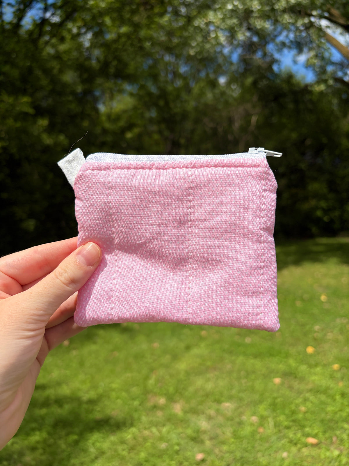 A small pink fabric zip pouch with a white zipper and a small white tag, held by a hand against a blurred green grass and tree background.
