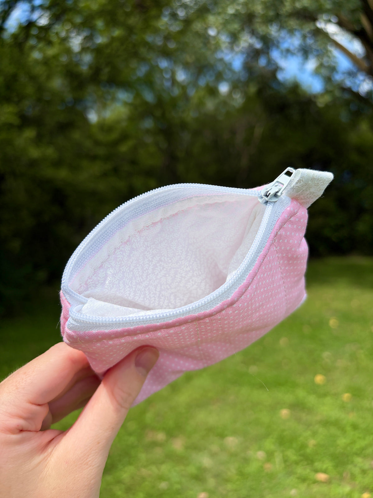 A small pink zippered pouch with a white interior, held in a hand against a blurred green outdoor background.