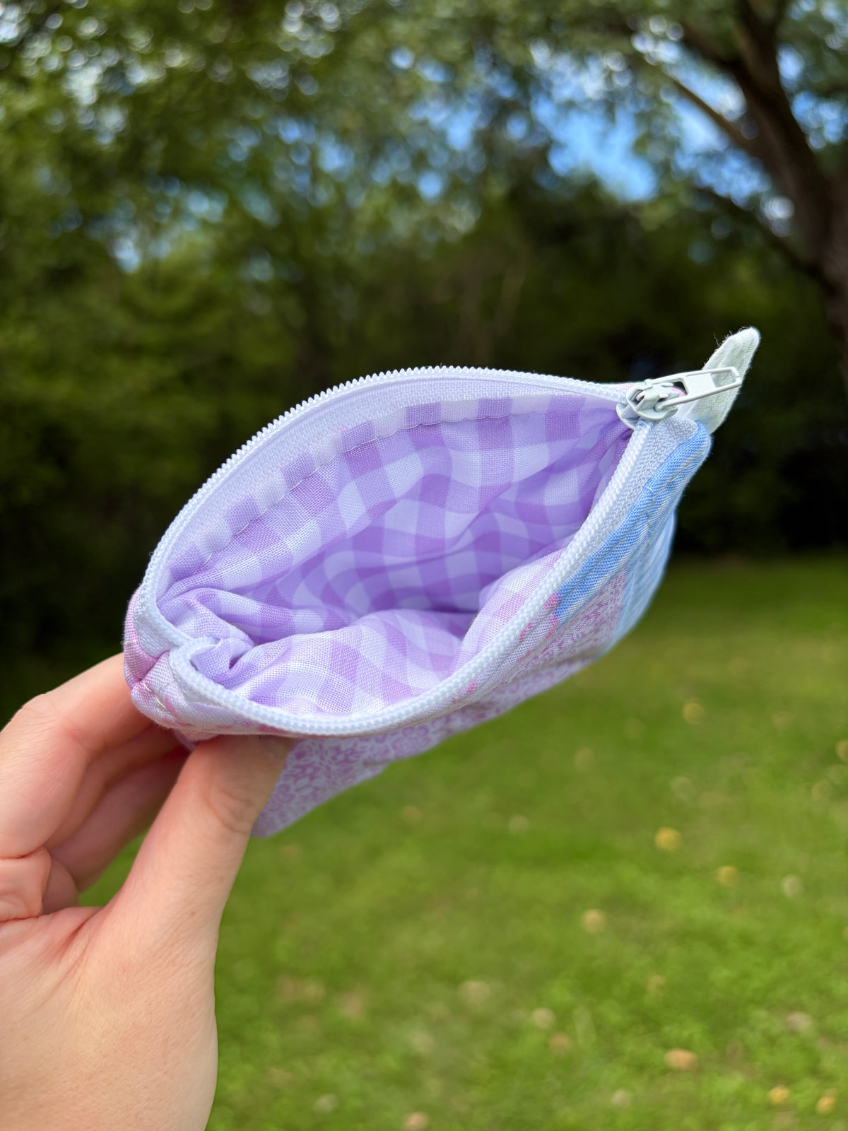 A hand holding an open zippered pouch with a lavender and white gingham pattern, set against a blurred green grassy background with trees.