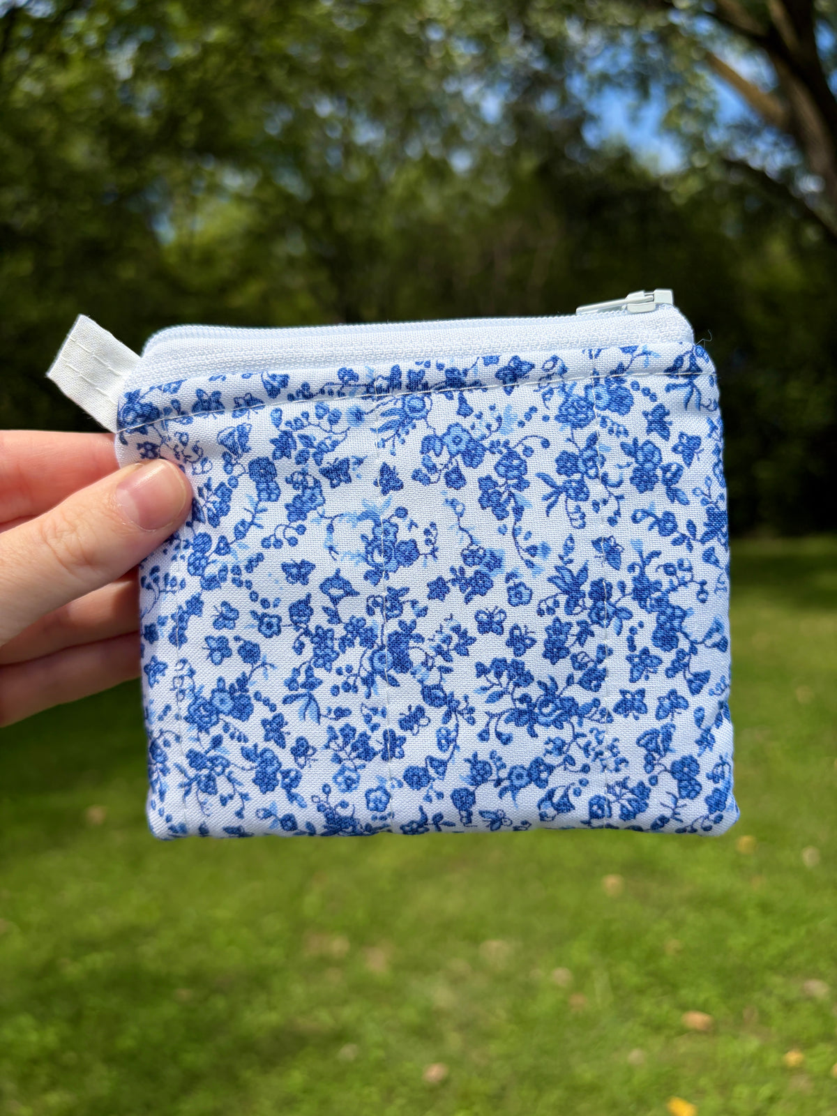 A hand holding a small zippered pouch with a blue and white floral toile de Jouy pattern, set against a blurred green grass and tree background.
