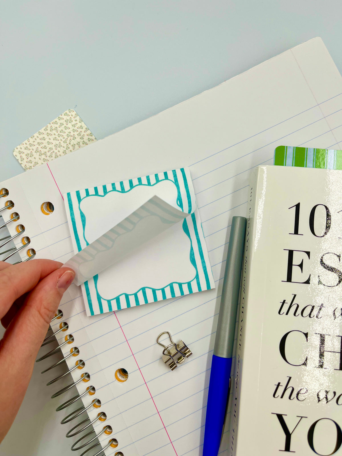 Hand holding a teal striped blank square sticky note on a lined notebook