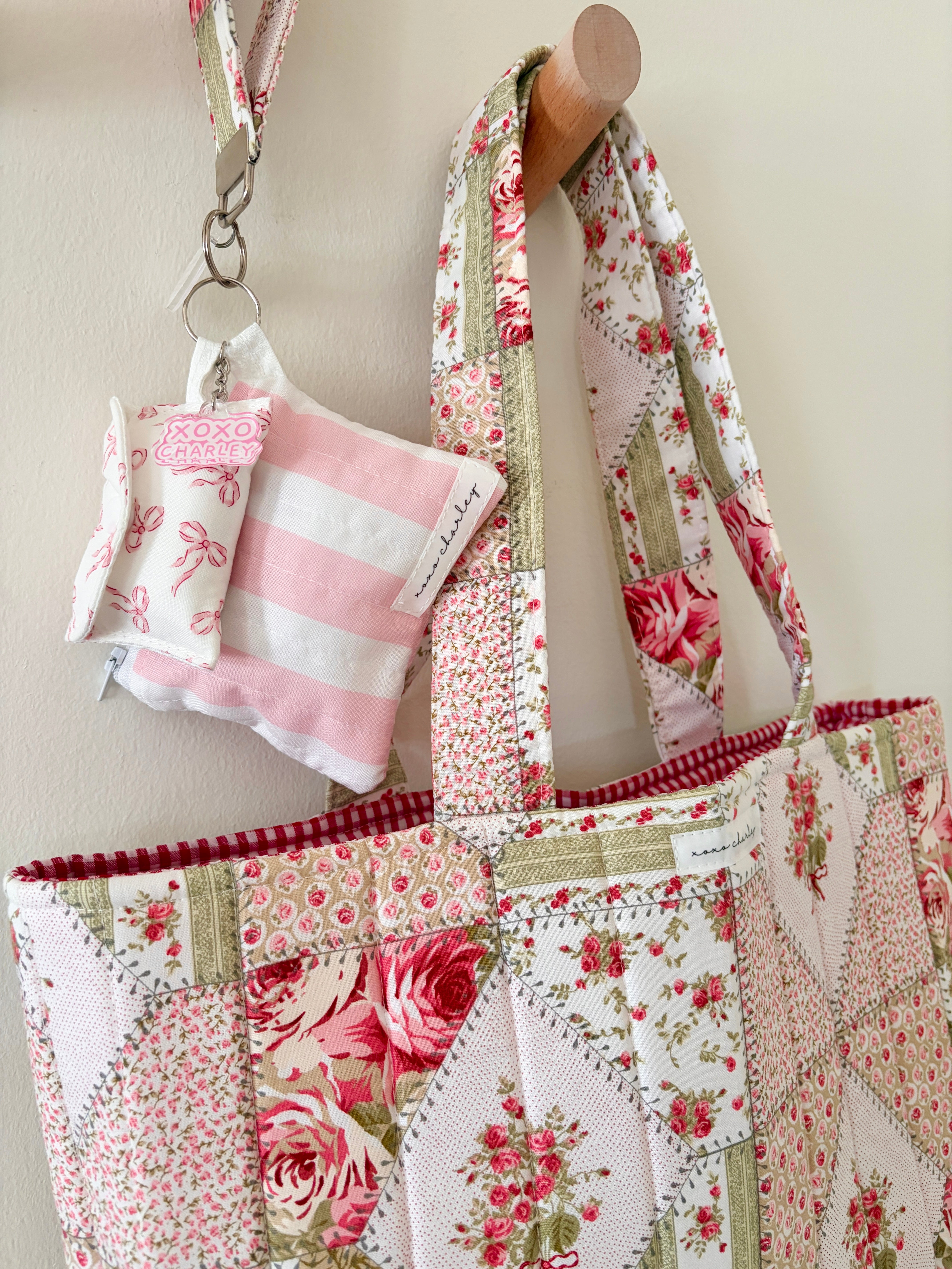 A patchwork quilted tote bag with floral and striped fabric panels, hanging from a wooden peg on a beige wall. A matching pink and white striped fabric pouch hangs from a key ring attached to the bag.