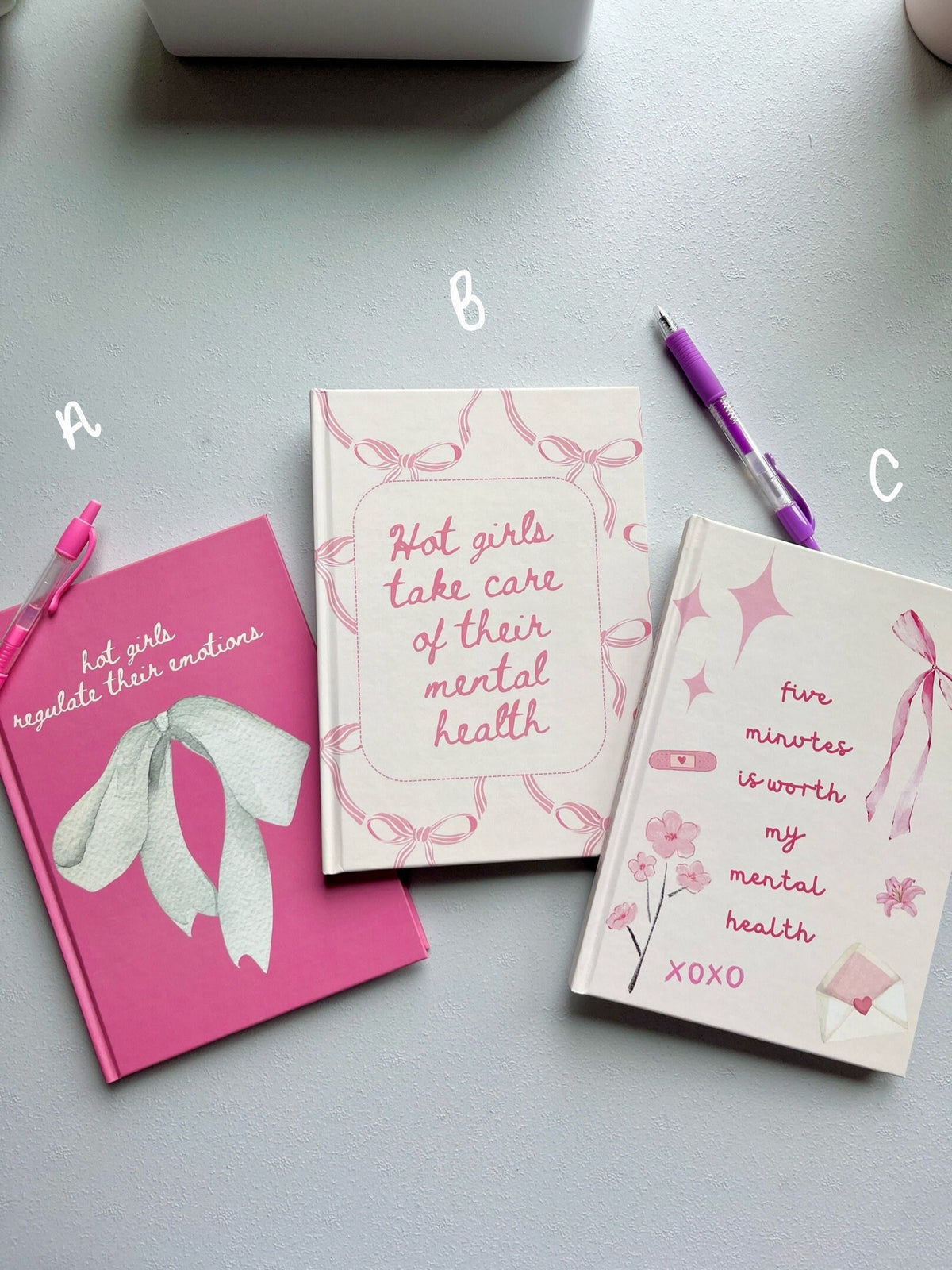 Mental health journals with pink and white covers, bow pattern, plus purple pen